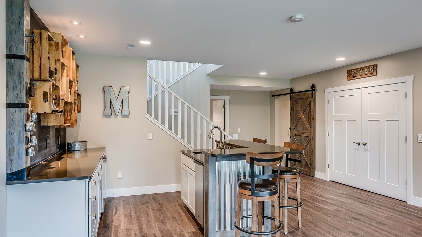 BASEMENT (5) Modern basement kitchen with a metal island, two bar stools, wooden shelves, double doors, and a sliding barn door beside a staircase.