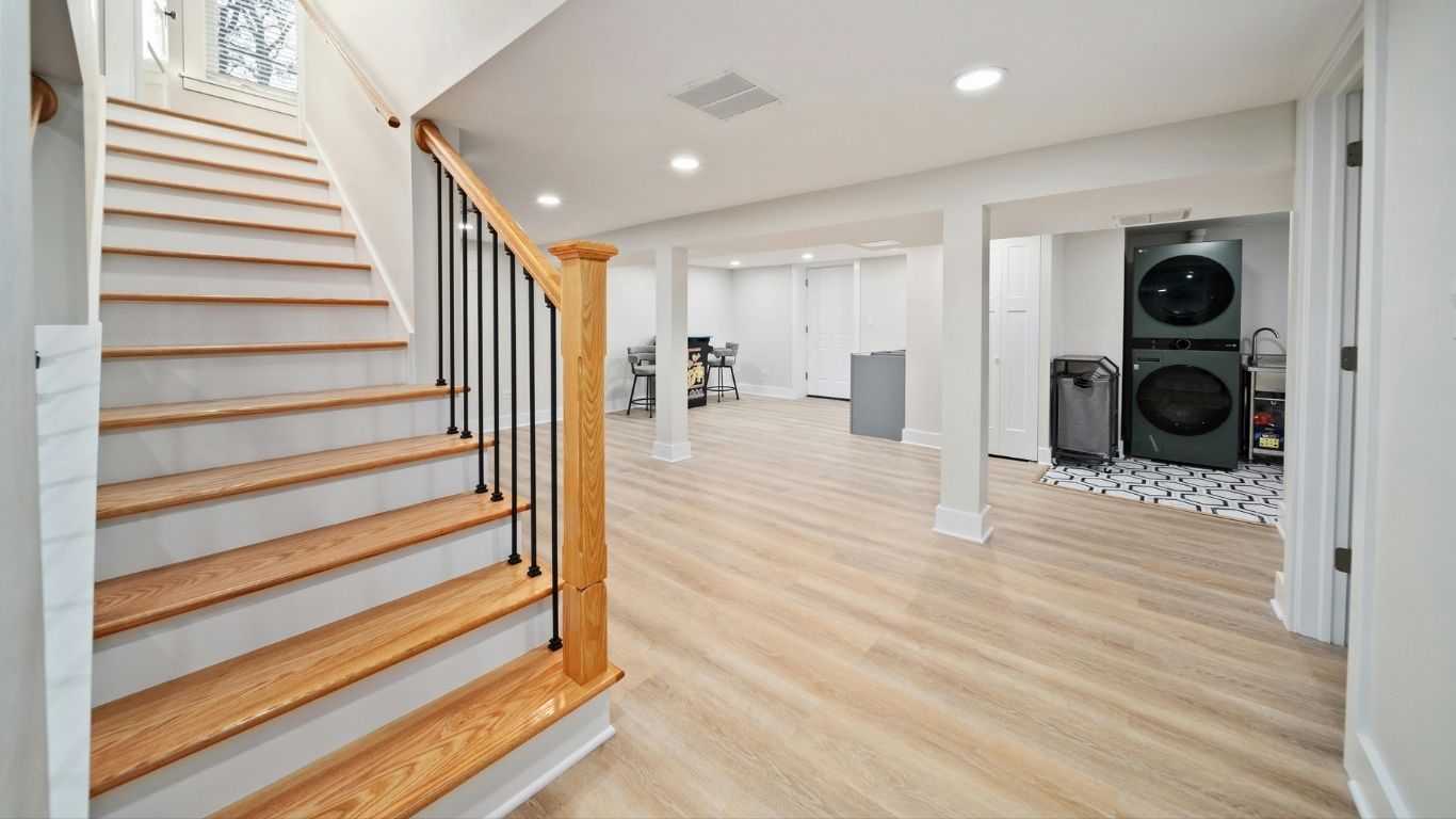 BASEMENT (7) A modern finished basement with light wood flooring, a wooden staircase, recessed lighting, a laundry area with stacked washer and dryer, and an open space with a table and chairs.