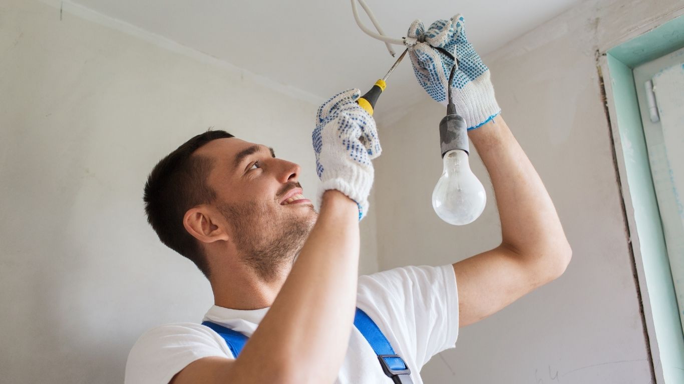 ELECTRICAL (4) Electrical Handyman - Lighting Handyman - Man wearing gloves and overalls fixes electrical wiring on the ceiling, holding a screwdriver near a hanging light bulb in a room with light-coloured walls.