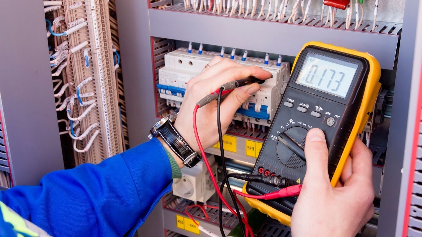 ELECTRICAL A person uses a digital multimeter to measure voltage inside an electrical control panel with various wires and circuit breakers.
