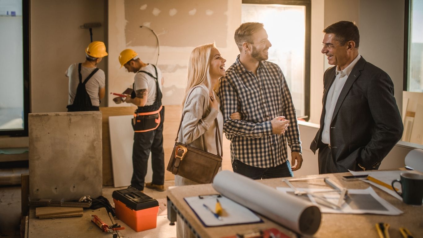 contractor talking (3) A couple speak with a man in a suit at a construction site, while two workers in hard hats work in the background. Building materials and plans are visible.