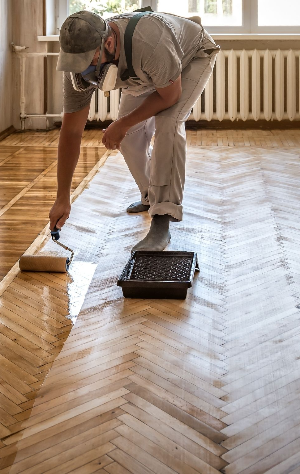 flooring (5) copy A person wearing a mask and cap applies varnish to a wooden parquet floor using a roller in a bright room with large windows and a radiator. - The Handyman Services York Region & Simcoe Region
