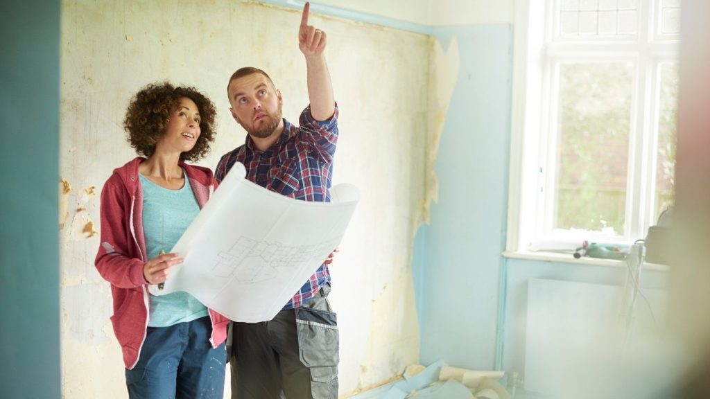 kitchen contractor (3) Two people stand in a partially renovated room, examining a blueprint; one person points towards the ceiling while the other looks in the same direction.