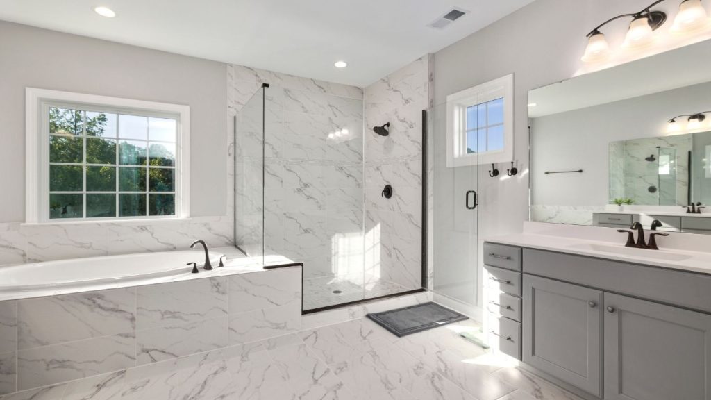 Budget-friendly bathroom remodeling. Modern bathroom with a large bathtub, glass-enclosed shower, grey vanity, wall mirror, and marble-style tiles throughout. Natural light enters through two windows. - The Handyman Services York Region & Simcoe Region