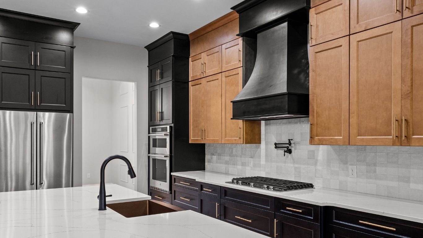 Refreshing kitchens and bathrooms: painting cabinets or replacing cabinet doors. Modern kitchen with black and wood cabinets, stainless steel appliances, a large black cooker hood, white worktops, a black sink tap, and under-cabinet lighting. - The Handyman Services York Region & Simcoe Region