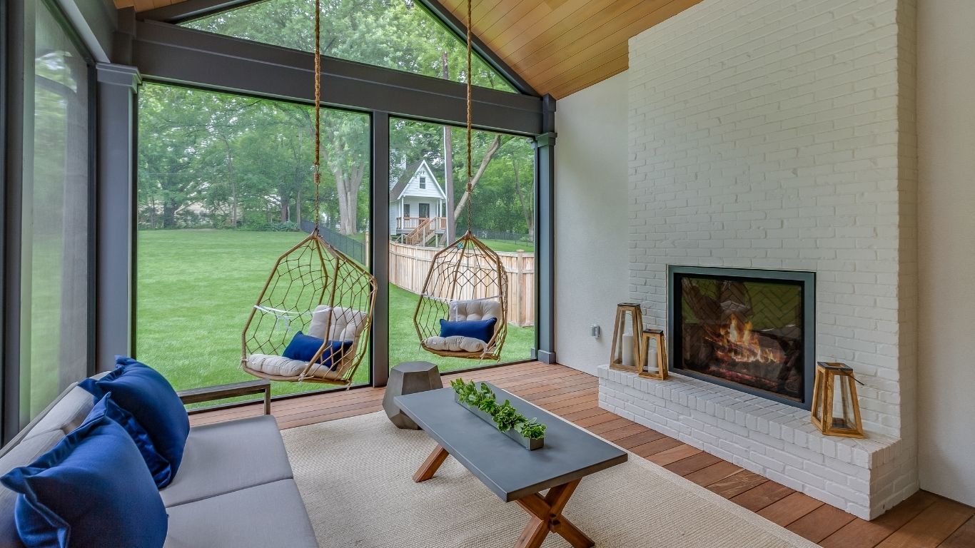 A screened-in veranda with two hanging chairs, a cushioned sofa, a coffee table with plants, and a white brick fireplace overlooking a green garden. - The Handyman Services York Region & Simcoe Region