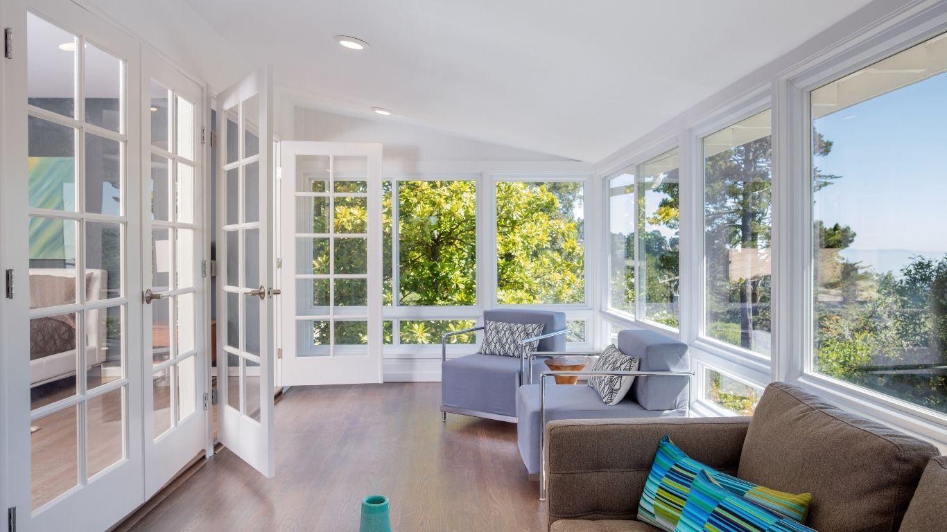 Bright sunroom with large windows, two armchairs, a sofa, and a side table, overlooking trees and outdoor scenery. French doors open to an adjoining room.