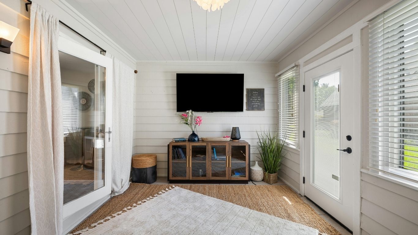 A bright sunroom with a TV mounted on the wall, a wooden cupboard, indoor plants, a rug, and large windows with blinds allowing natural light.
