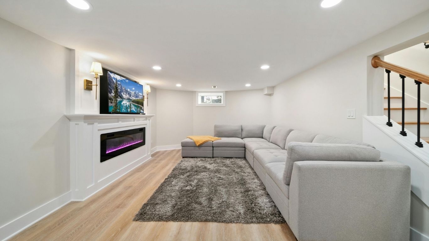 Basement living room with a grey corner sofa, rug, wall-mounted TV above a fireplace, and wooden stairs with black bannister.