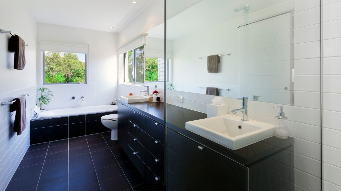Modern bathroom renovation with black tiled floor, white walls, a bath by two windows, a toilet, and a double sink vanity with a large mirror.