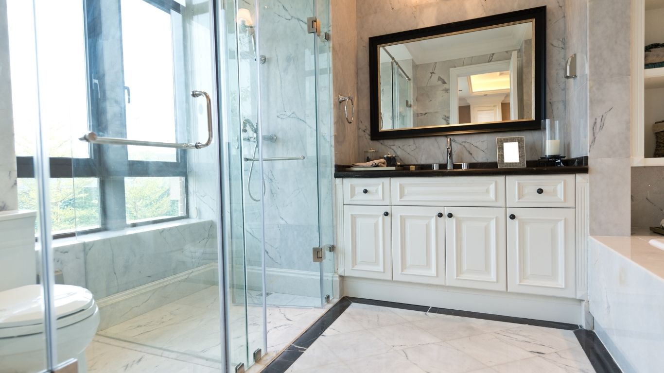 Modern bathroom with glass shower enclosure, white cupboards, large mirror, marble floors and walls, and a window letting in natural light.