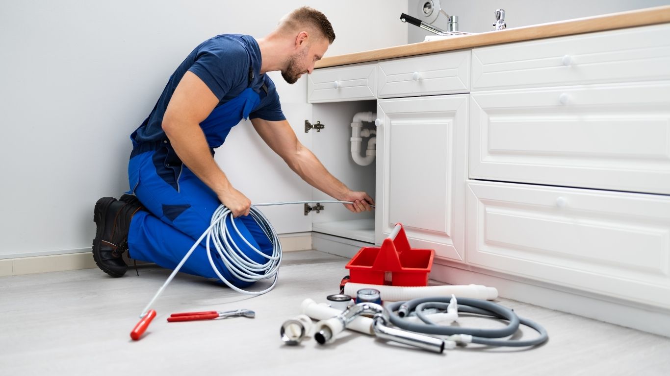 A plumber in blue overalls kneels by a kitchen sink, holding a pipe and hose, with plumbing tools and supplies on the floor nearby.