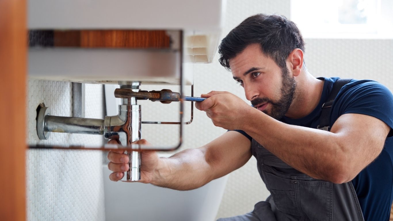 Plumbing and Faucet Repair Simcoe and York Regions - The Handyman Services - A man in overalls uses a spanner to repair plumbing under a sink in a bathroom.