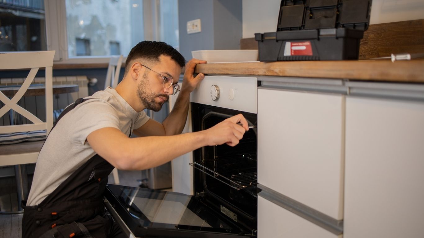 General Home Repairs Handyman - A man in work overalls is kneeling and repairing a built-in oven in a modern kitchen, using a tool. A toolbox is on the worktop above him. - The Handyman Services Aurora