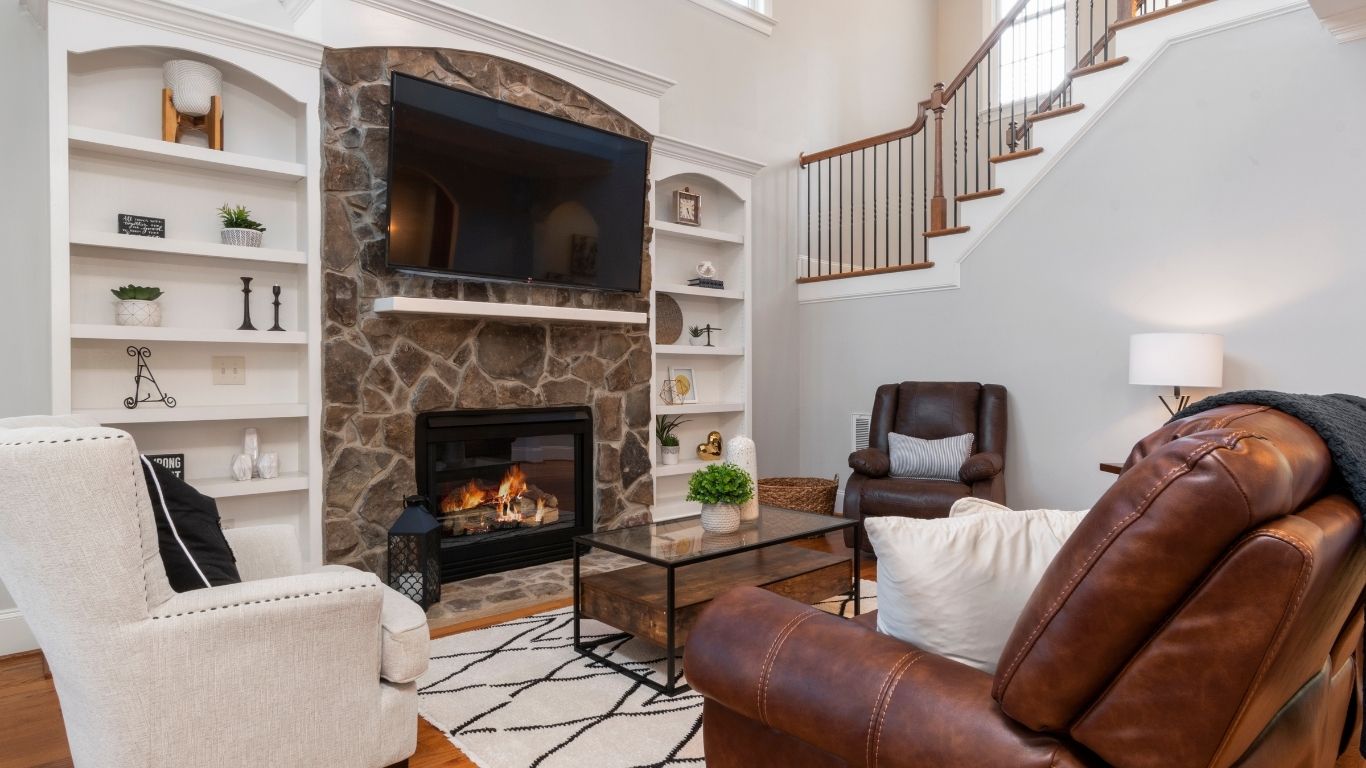 A living room with a stone fireplace, wall-mounted TV, two armchairs, a leather sofa, glass coffee table, and built-in shelves, with stairs and a lamp in the background. - The Handyman Services Aurora