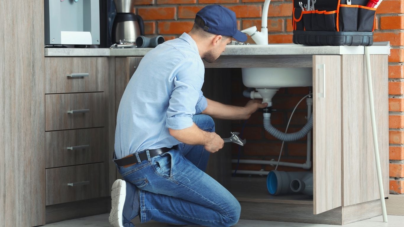 A plumber in a blue cap and shirt kneels in front of an open cupboard, repairing pipes under a kitchen sink with tools beside him. - The Handyman Services Barrie