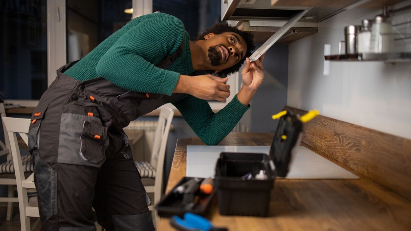 The Handyman Services East Gwillimbury - general repairs A man in work overalls repairs a kitchen cupboard using tools from a black toolbox on the worktop. - The Handyman Services East Gwillimbury