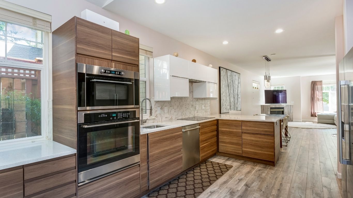 Modern kitchen with wooden cabinetry, built-in stainless steel appliances, white worktops, tiled splashback, and an open view to the dining and living area with large windows. - The Handyman Services Richmond Hill