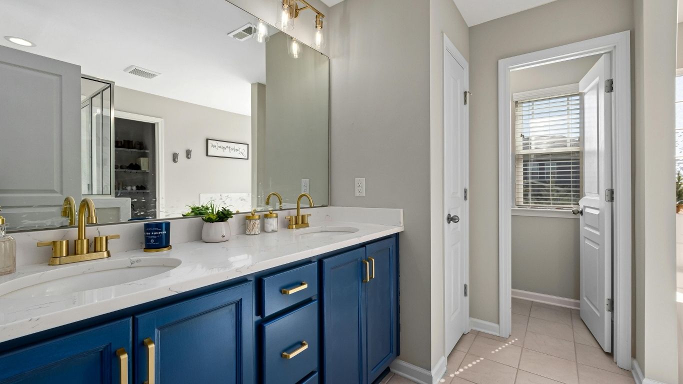 Modern bathroom with double sink vanity featuring blue cupboards, gold fittings, large mirror, and view into a sunlit adjoining room with tiled flooring. - The Handyman Services East Gwillimbury