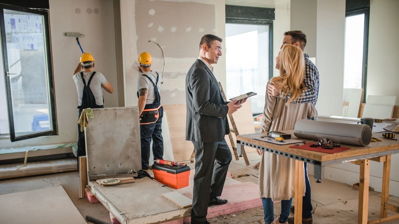 A man in a suit talks to a couple inside a building undergoing refurbishment while two construction workers work in the background.