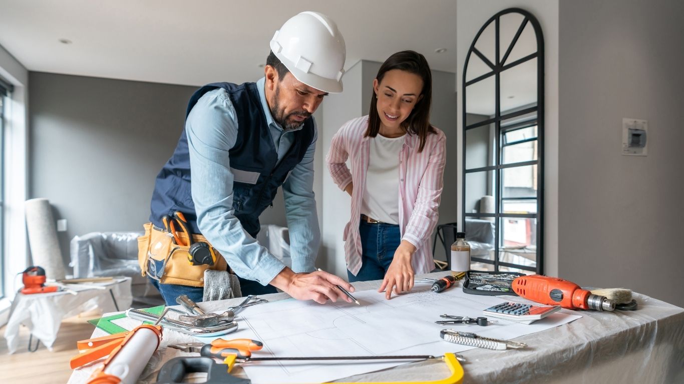 Bathroom Renovation Contractor A man in a construction helmet and a woman review blueprints on a table covered with tools in a modern, unfinished room.