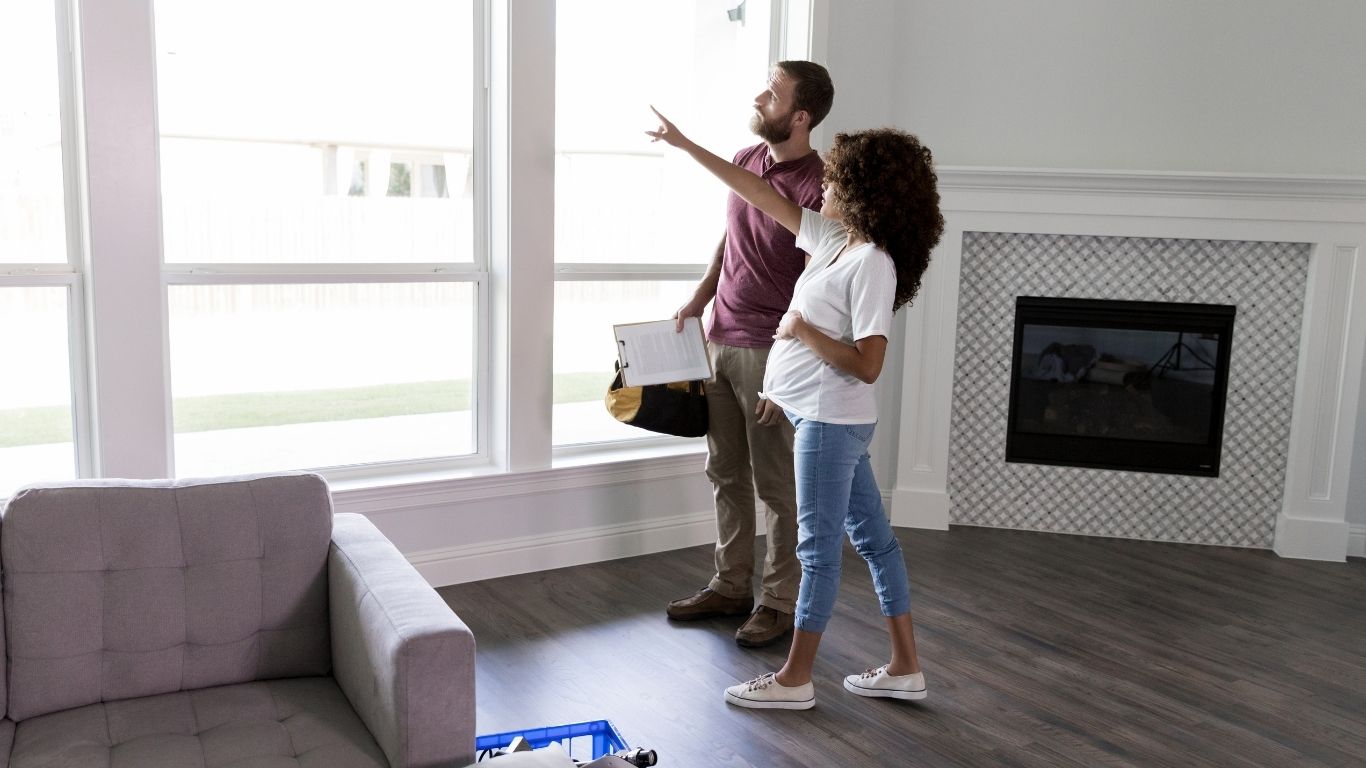 Two people stand in an unfurnished living room with wooden floors and large windows, discussing the space. One is pointing towards the window while the other holds a clipboard and bag.