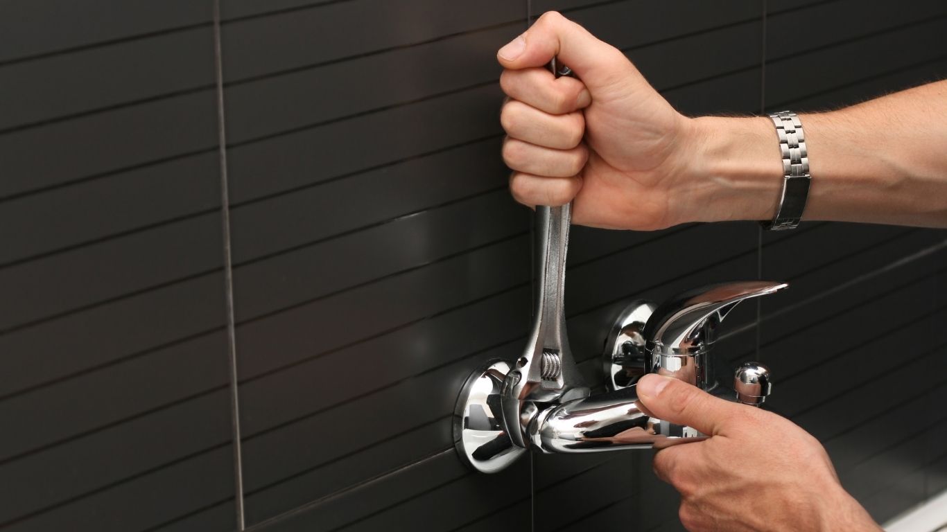 Handyman Services - Plumbing - A person uses a spanner to tighten or adjust a wall-mounted chrome tap against a dark tiled wall.