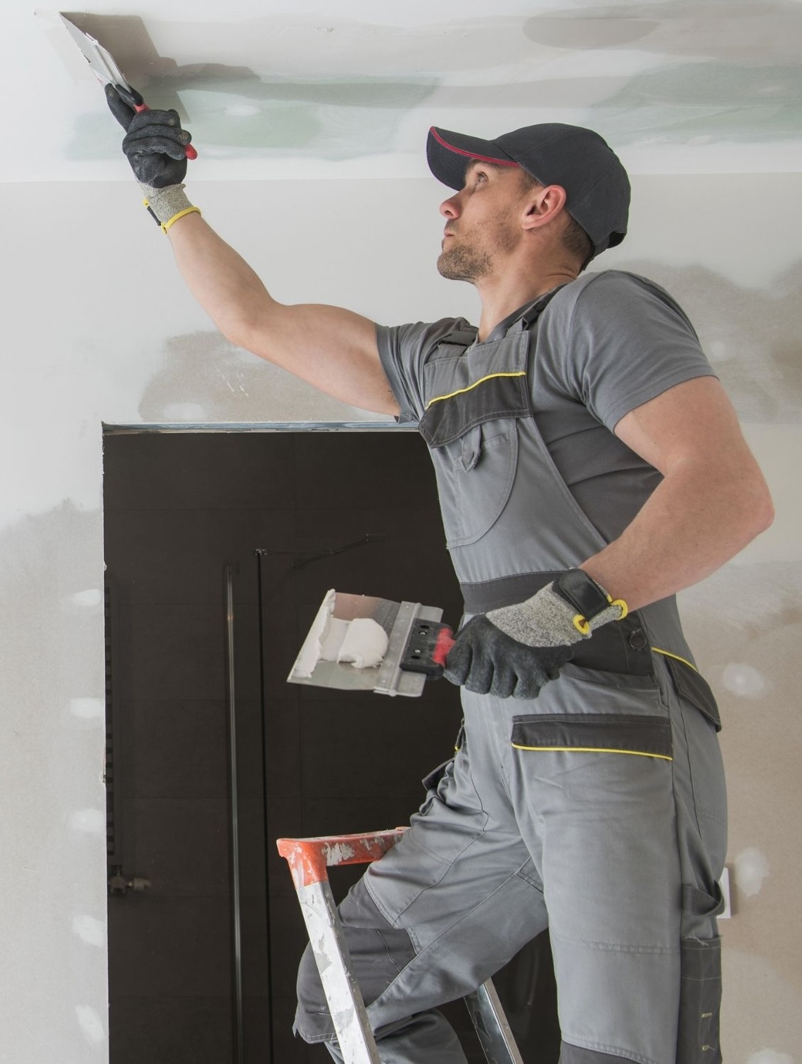 A person in grey work overalls stands on a ladder, applying plaster to a ceiling with a trowel, holding additional tools in the other hand. - The Handyman Services York Region & Simcoe Region