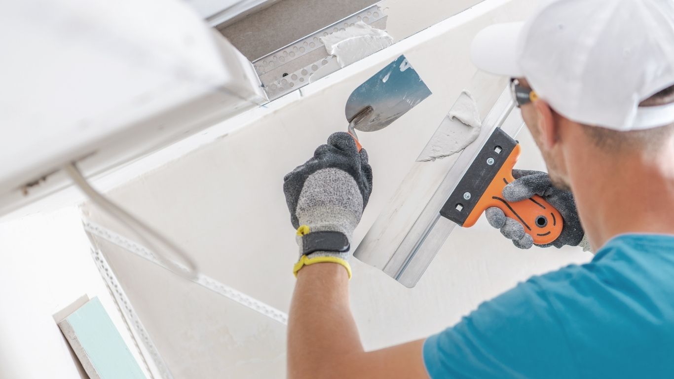 Person wearing gloves and a cap applies jointing compound to a ceiling with a plasterboard knife and trowel. - The Handyman Services York Region & Simcoe Region