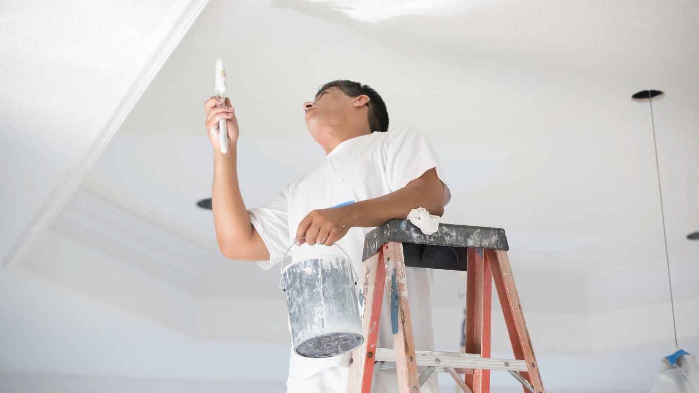 A man standing on a ladder paints a white ceiling with a brush, holding a paint tin in his other hand. - The Handyman Services York Region & Simcoe Region